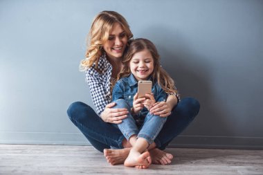 Sevimli küçük kız ve güzel genç annesiyle birlikte yere oturmak, bir akıllı telefon kullanarak ve, gri arka plan üzerinde gülümseyen