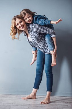 Sevimli küçük kız ve güzel genç annesini kameraya bakarak ve, gri arka plan üzerinde gülümseyen. Kız pickaback oturuyor