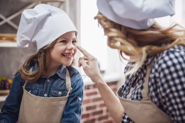 Cute little girl and her beautiful mom in aprons and chef hats are touching noses and smiling while kneading the dough in the kitchen
