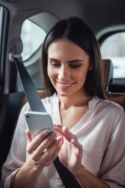 Güzel iş kadını bir akıllı telefon kullanarak ve arabanın arka koltukta otururken gülümseyen