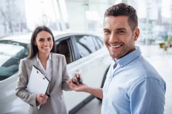 Visiting car dealership. Handsome man and beautiful sales manager are holding car key, looking at camera and smiling