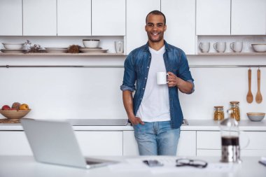 Yakışıklı iş adamı kahve içiyor, kameraya bakıyor ve mutfakta dururken gülümsüyor.