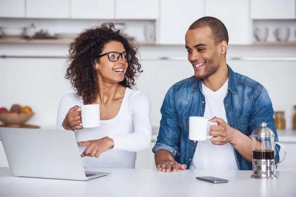 Beautiful couple is drinking coffee, talking and smiling while using a laptop in kitchen