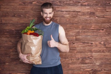 Spor giyim yakışıklı genç sakallı adam Ok işaret gösteren ve gülümseyen, ahşap arka plan üzerinde sağlıklı gıda, bir alışveriş torbası tutuyor