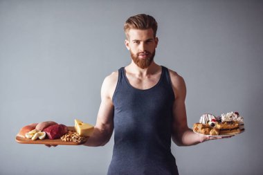 Yakışıklı genç sakallı sporcu sağlıklı gıda ile ahşap tepsi ve bir plaka ile kek, gri arka plan üzerinde tutuyor