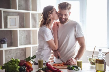 Güzel genç çift konuşmak ve sağlıklı gıda evde mutfakta yemek yaparken gülümseyerek. Kız erkek arkadaşı yanak içinde öpüşme