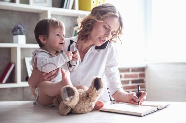 Güzel iş anne cep telefonda konuşurken ve onun şirin bebek çocuk evde vakit sırasında notlar almak