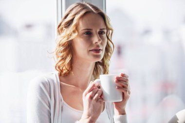 Güzel kadın bir fincan tutarak ve uzakta pencere pervazına üzerinde evde otururken arıyorsunuz