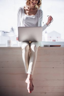 Güzel kadın bir dizüstü bilgisayar kullanarak ve pencere pervazına üzerinde evde otururken gülümseyen