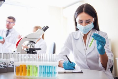 Eldiven ve maske alanlarındaki güzel tıp doktoru laboratuarda test tüpleri ile çalışırken notları yapıyor