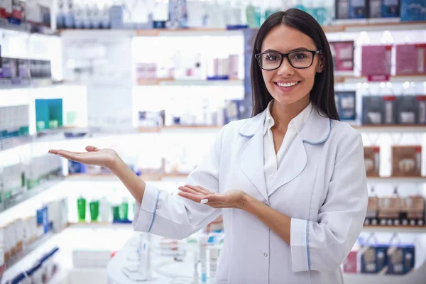 Beautiful female pharmacist is showing shelves with medicine, looking at camera and smiling while working in pharmacy