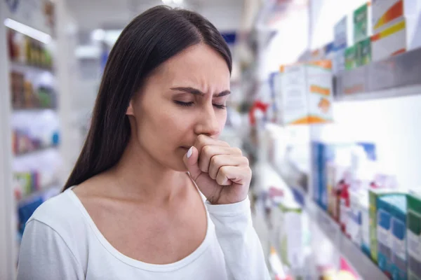 Beautiful young woman is coughing while choosing a remedy at the pharmacy