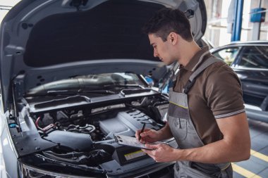 Üniformalı yakışıklı genç otomatik mekanik Oto Servisi arabaya incelenirken notlar yapıyor