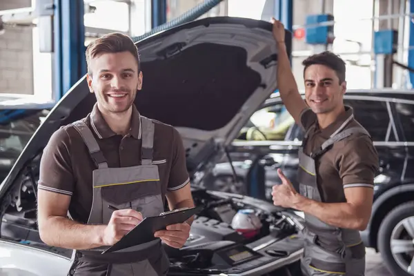 Üniformalı yakışıklı genç otomatik mekanik kameraya bakarak ve oto Servisi arabaya incelenirken gülümseyen