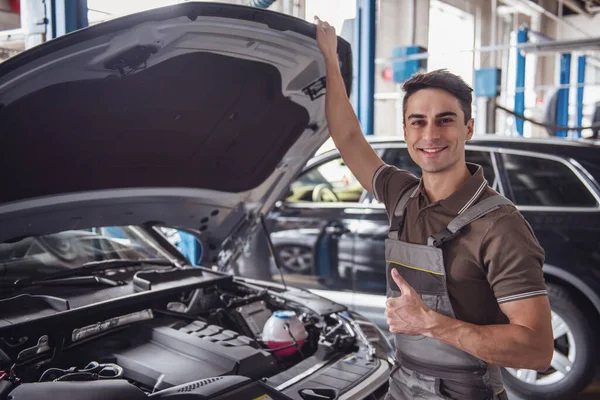 Handsome young auto mechanic in uniform is showing Ok sign, looking at camera and smiling while examining car in auto service