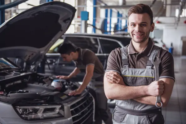 At the auto service. Handsome young auto mechanic in uniform is looking at camera and smiling while his colleague is examining car