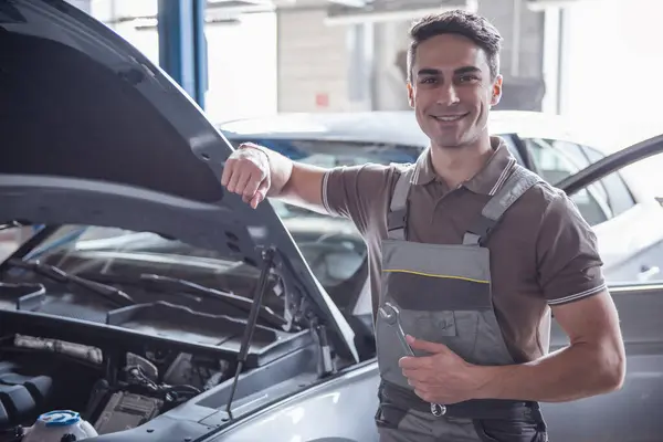 Attractive young auto mechanic in uniform is showing Ok sign, looking at camera and smiling while standing in auto service