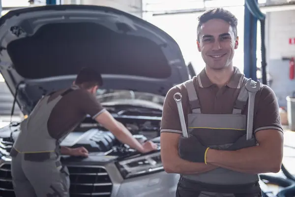 At the auto service. Handsome young auto mechanic in uniform is holding tools, looking at camera and smiling while his colleague is examining car