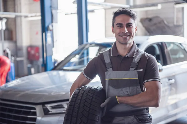 At the auto service. Handsome young auto mechanic in uniform is holding a tire, looking at camera and smiling