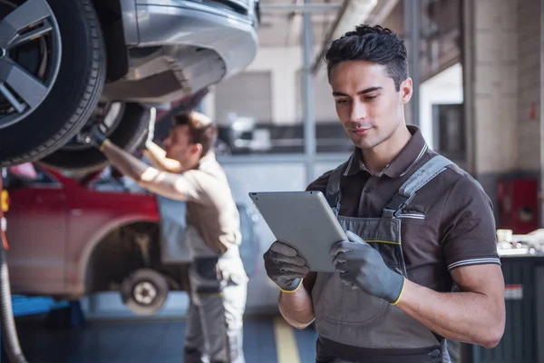 At the auto service. Handsome young auto mechanic in uniform is using a digital tablet while his colleague is examining car