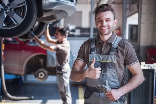 At the auto service. Handsome young auto mechanic in uniform is showing Ok sign, looking at camera and smiling while his colleague is examining car