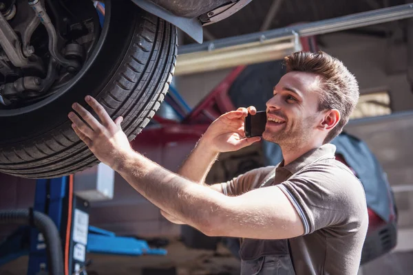 At the auto service. Handsome young auto mechanic in uniform is talking on the mobile phone and smiling while examining car