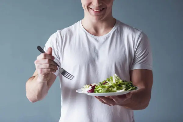 Handsome guy is smiling while holding a plate with salad, on gray background