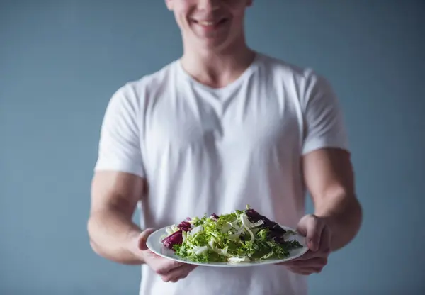 Handsome guy is smiling while holding a plate with salad, on gray background