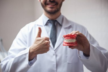 Yakışıklı genç diş hekimi Beyaz önlük bir çene holding, Ok işaret gösteren ve ofisinde dururken gülümseyen