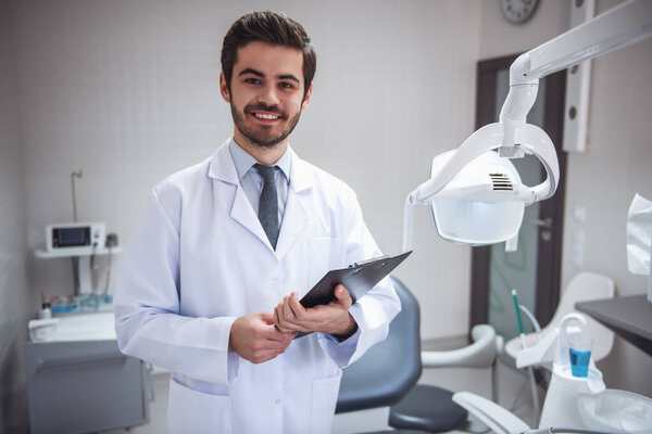 Handsome young dentist in white coat is holding a folder, looking at camera and smiling while standing in his office