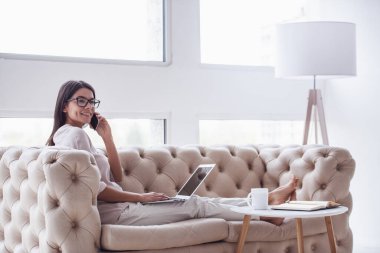 Ev ofis. Oturma odasında kanepede oturan başarılı genç güzel kadın. dizüstü bilgisayar kullanarak ve telefonda konuşurken