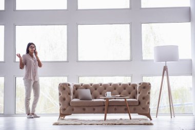 Ev ofis. Oturma odasında büyük bir pencere yakınında duran ve telefonda konuşurken başarılı genç güzel kadın