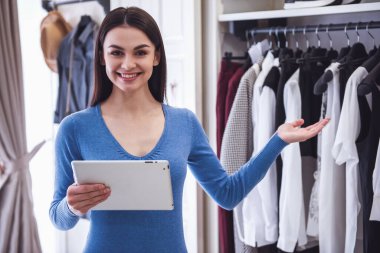Güzel kadın tezgâhtar dijital tablet, giysiler için işaret, butik dururken gülümseyen ve kameraya bakarak kullanma