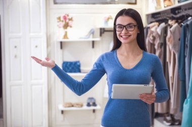 Güzel kadın tezgâhtar dijital tablet, giysiler için işaret, butik dururken gülümseyen ve kameraya bakarak kullanma