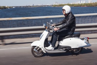 Deri ceket ve kask adam Şehir Köprüsü aracılığıyla scooter sürme ve gülümseyen