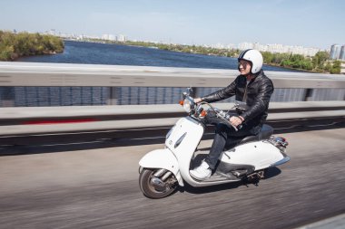 Deri ceket ve kask adam Şehir Köprüsü aracılığıyla scooter sürme ve gülümseyen