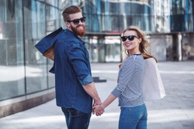 Güzel genç çift güneş gözlüğü ve poşetler el ele tutuşarak, kameraya bakarak ve yürürken gülümseyen aşağı sokak