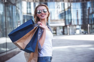 Güneş gözlüklü güzel kız alışveriş torbaları holding, kameraya bakıyor ve sokakta yürürken gülümseyen