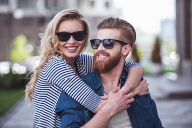 Sarılma, kameraya bakıyor ve açık havada duran süre gülümseyen güneş gözlüğü güzel genç Çift