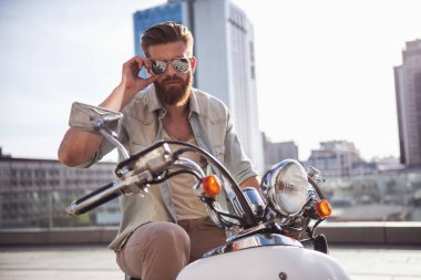 Güneş gözlüklü yakışıklı sakallı adam onun scooter üzerinde otururken kameraya bakıyor