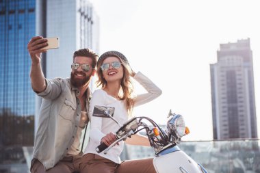 Güzel genç çift güneş gözlüklü bir akıllı telefon kullanarak ve bir scooter açık havada üzerinde otururken gülümseyen selfie yapıyor