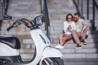 Güzel genç bir çift bir akıllı telefon kullanarak ve scooter ön planda açık havada, merdivenlerde otururken gülümseyen