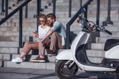Güzel genç bir çift bir akıllı telefon kullanarak ve scooter ön planda açık havada, merdivenlerde otururken gülümseyen