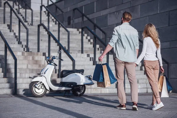 Güzel genç çiftin alışveriş çantaları taşırken ve onların scooter için gelirken elele arkadan görünüm