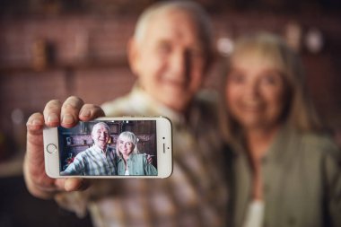 Güzel üst düzey çift sarılma, bir akıllı telefon kullanarak ve mutfakta dururken gülümseyen selfie yapıyor. Odakta telefon
