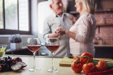 Güzel üst düzey çift dans ve birlikte mutfakta yemek yaparken gülümseyerek. Bardak şarap ön planda