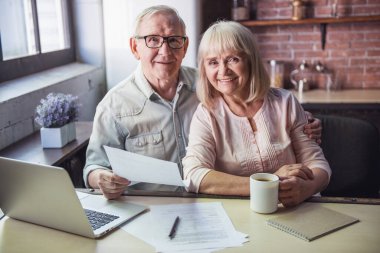 Güzel üst düzey iş çift belgeleri eğitim, bir dizüstü bilgisayar kullanarak, kameraya bakıyor ve mutfakta çalışırken gülümseyen