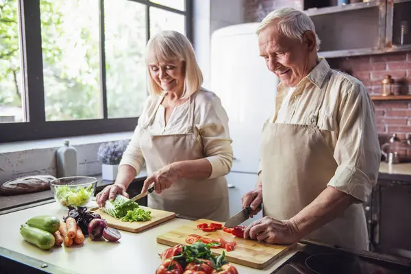 Beautiful senior couple in aprons is talking and smiling while cooking together in kitchen