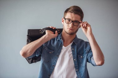 Rahat kıyafetler ve gözlük yakışıklı genç adam kameraya bakıyor ve gülümsüyor, gri arka plan üzerinde şık bir çanta tutuyor