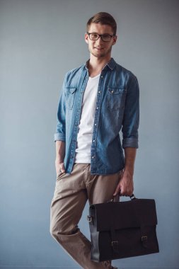 Yakışıklı genç işadamı rahat kıyafetler, gözlük ve şık bir çanta ile kameraya bakarak ve gri arka plan üzerinde gülümseyen